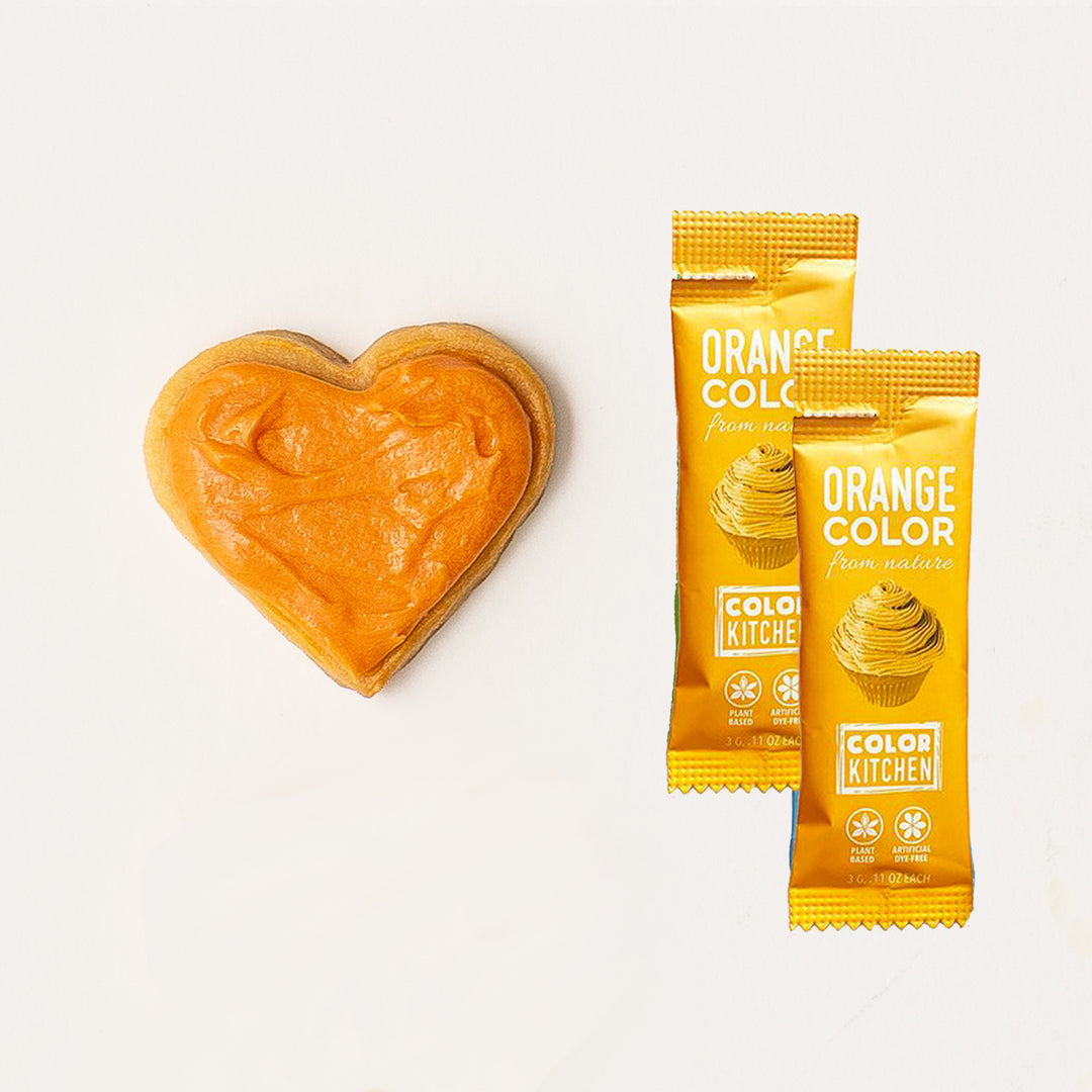 Two packets of natural orange powdered food color next to a heart-shaped sample of dye-free colored orange frosting.