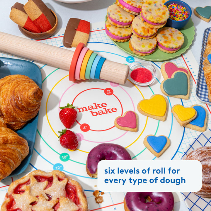 Baking setup with cookies, pastries, and a rolling pin on a 'make bake' mat.