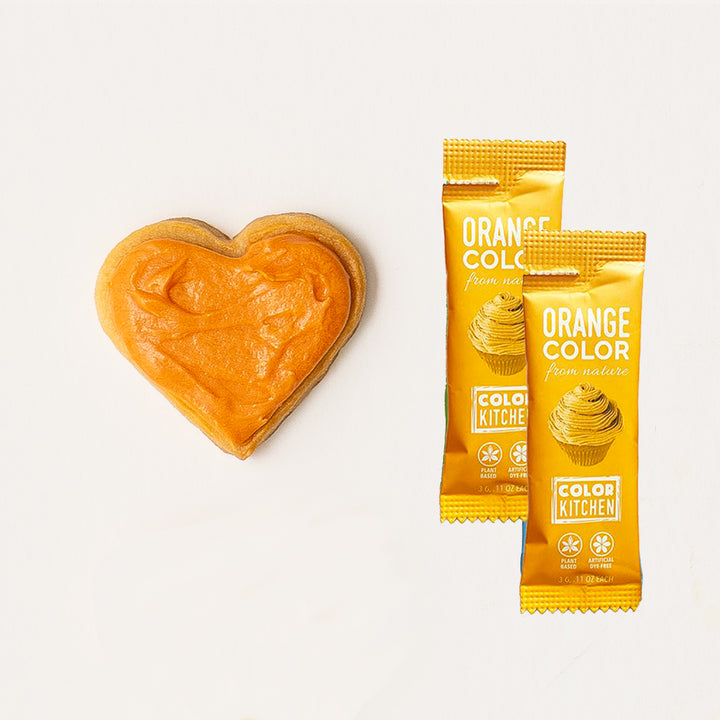 Two packets of natural orange powdered food color next to a heart-shaped sample of dye-free colored orange frosting.