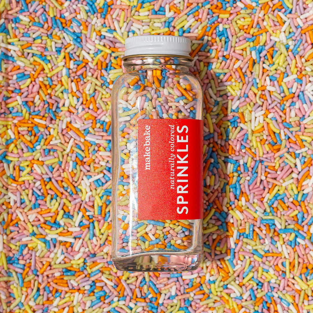 A clear glass jar filled with naturally colored rainbow sprinkles in pastel colors placed on a background covered with similar sprinkles, with a red 'Make Bake' label on the jar.
