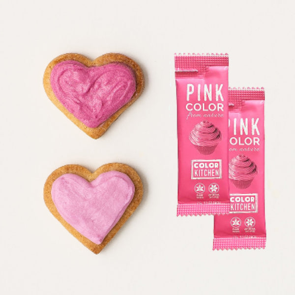 Two packets of natural pink food color next to heart-shaped cookies frosted with dye-free pink icing.