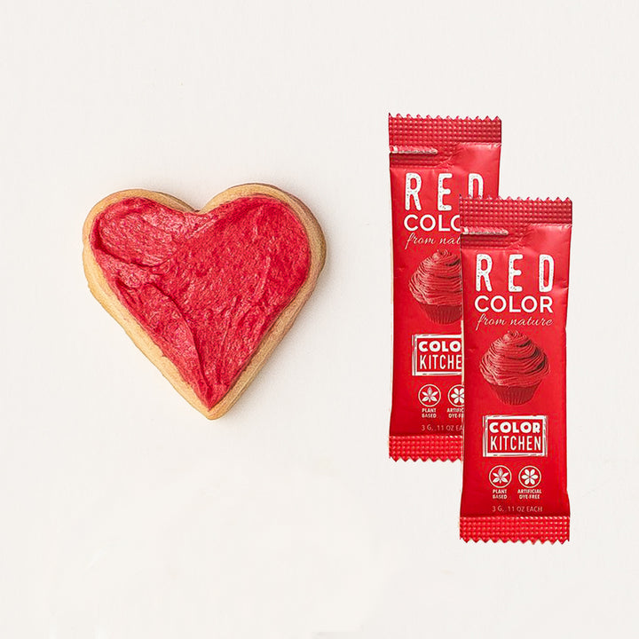 Two packets of natural red food coloring next to a heart-shaped cookie decorated with the dye-free red frosting.