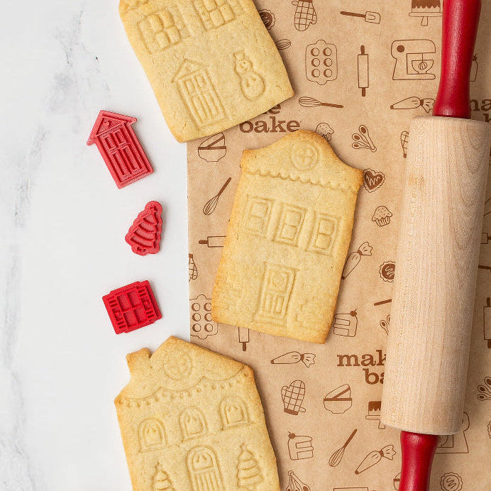 Decorative sugar cookies shaped like houses with a rolling pin and cookie stamps on parchment paper.