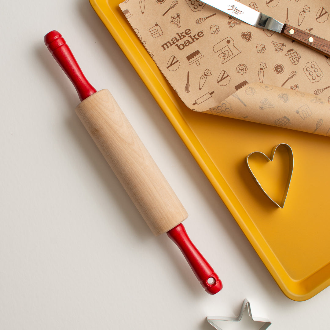 A wooden rolling pin with red handles lies on a yellow baking tray alongside a heart-shaped cookie cutter, brown patterned baking paper, and a metal spatula on a light neutral background.
