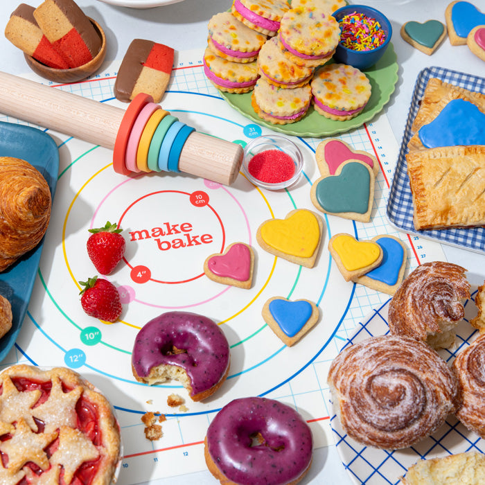 Assorted baked goods including heart cookies, blueberry donuts, cinnamon rolls, strawberry pie on a 'make bake' pastry mat and with Make Bake's rainbow roller adjustable rolling pin with guide rings in rainbow colors.