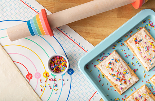 Large wooden rolling pin with adjustable rings used to make hand pies with natural, dye free rainbow sprinkles shown on a light blue baking sheet on a kitchen butcher block countertop.