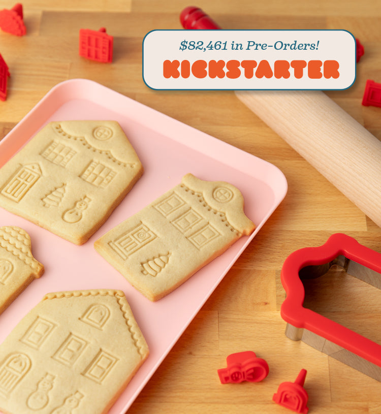 House-shaped cookies on a pink tray with cookie cutters on a wooden surface, text says "$82,461 in Pre-Order! on Kickstarter."
