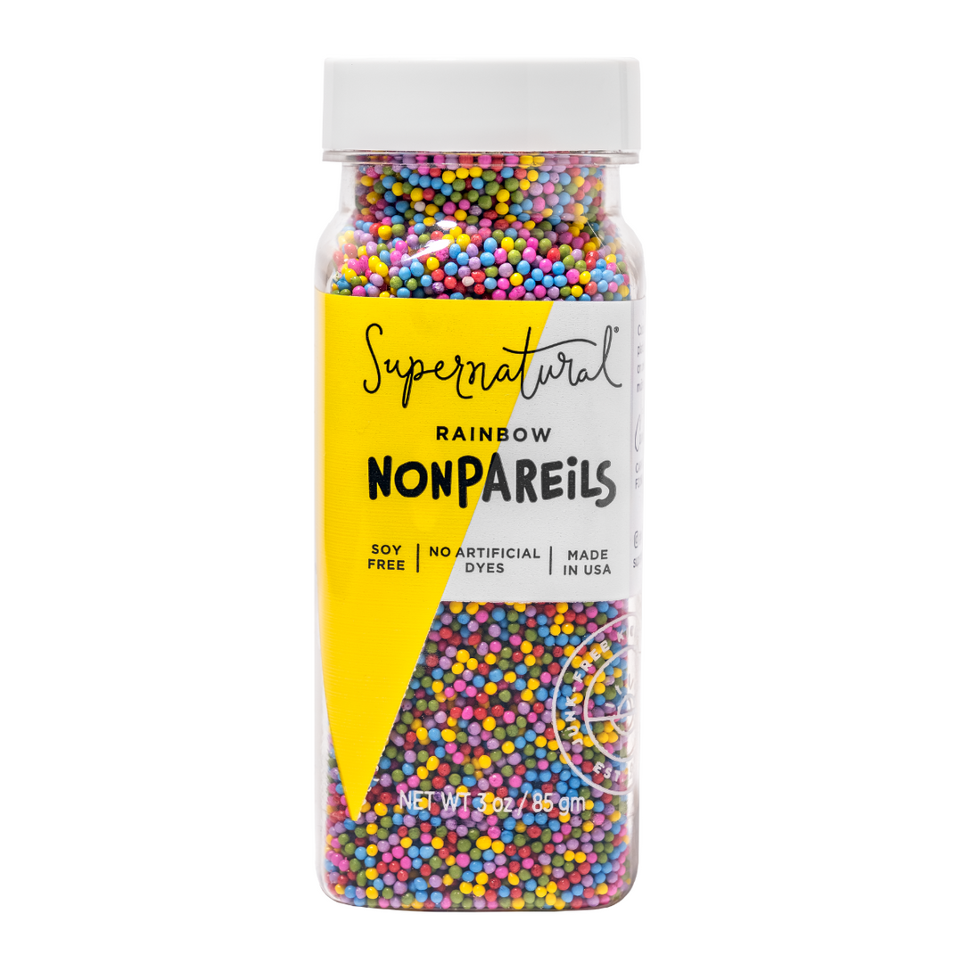 A jar of Supernatural brand dye-free rainbow nonpareils, with colorful sprinkles visible in a the clear bottle. The label is yellow with text detailing the product being soy-free, non-GMO, and with no artificial dyes.