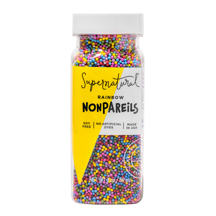 A jar of Supernatural brand dye-free rainbow nonpareils, with colorful sprinkles visible in a the clear bottle. The label is yellow with text detailing the product being soy-free, non-GMO, and with no artificial dyes.