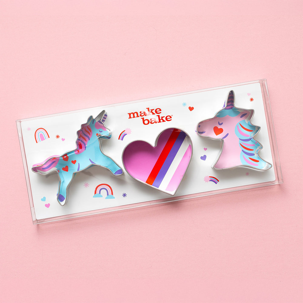 A set of three cookie cutters in the shapes of a unicorn and a heart with a rainbow background, packaged in a clear plastic box with 'Make Bake' branding.