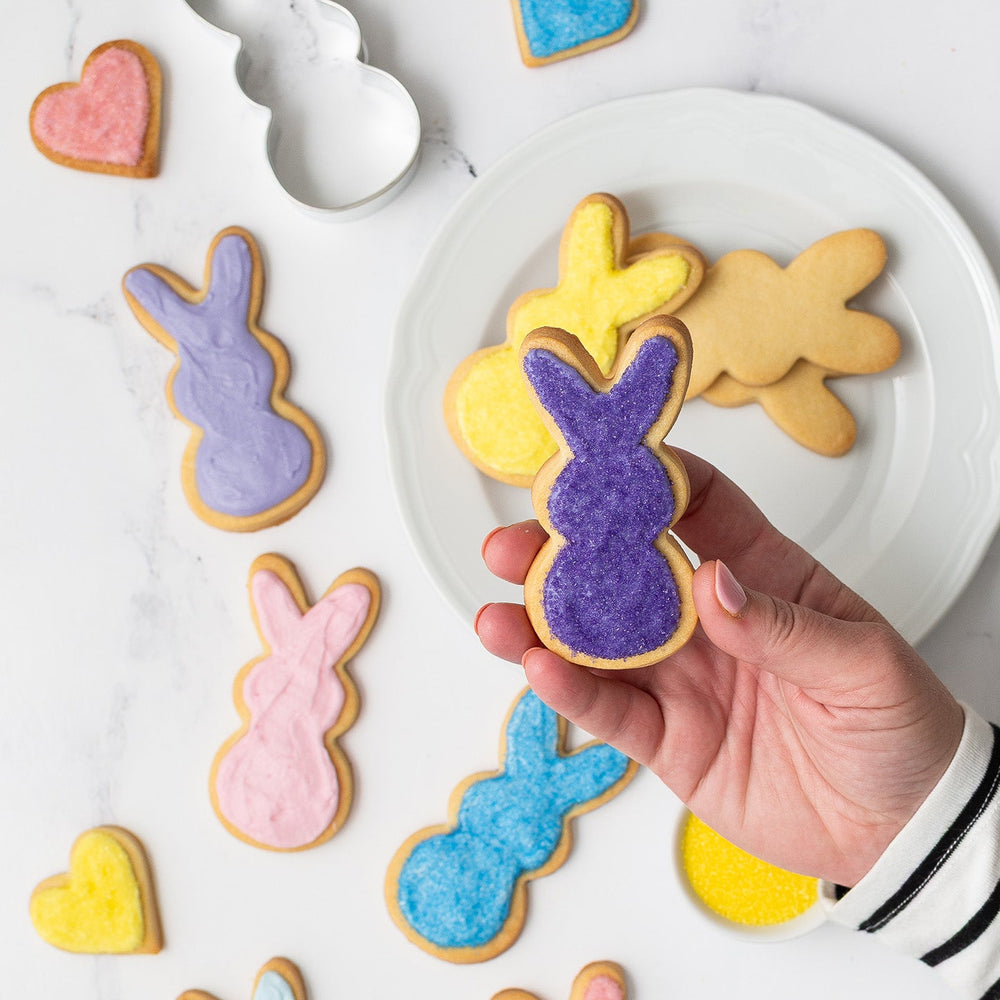 Colorful bunny-shaped cookies on a white plate with a hand holding a bunny-shaped cookie decorated with natural purple food color and sugars.