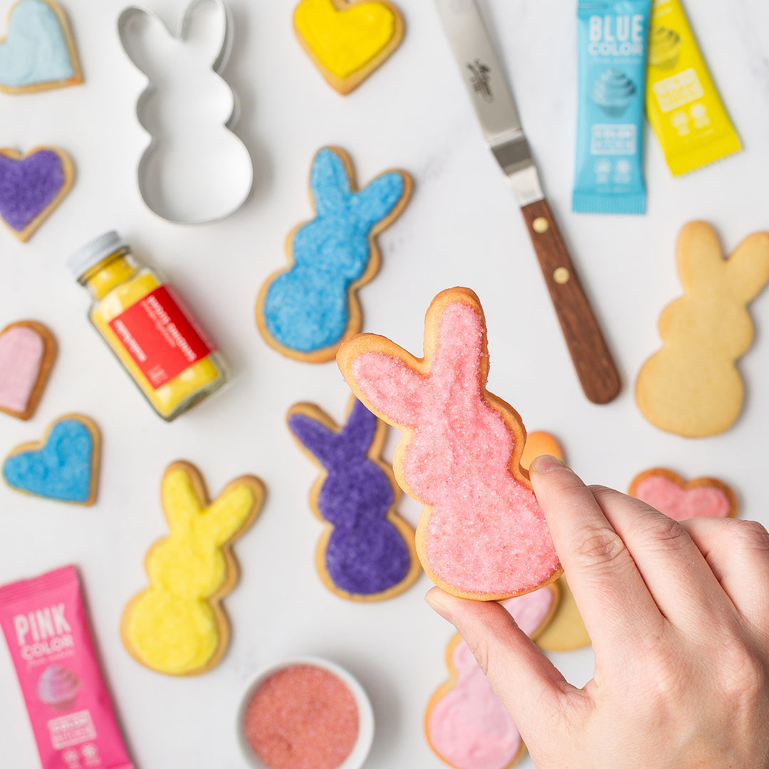 hand holding a bunny sugar cookie decorated with dye-free sanding sugar and more cookies in the background in  pink, purple, blue and yellow