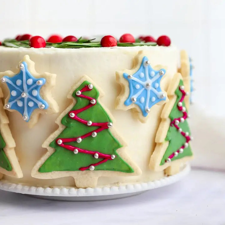 White round cake decorated with Christmas trees and snowflakes made with naturally colored frosting on a white plate and topped with cranberries and greenery