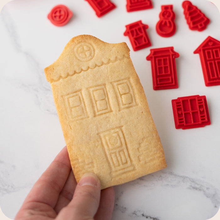 Hand holding a cookie shaped like a house with red cookie stamps on a marble surface.