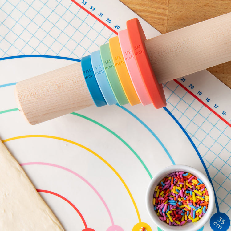 Wooden rolling pin with colorful guide bands on a baking sheet with rainbow design and sprinkles.