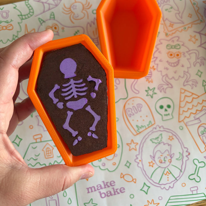 Picture of a hand holding a chocolate brownie in the shape of of a coffin with a purple skeleton stenciled on in frosting with a halloween themed paper table runner in the background