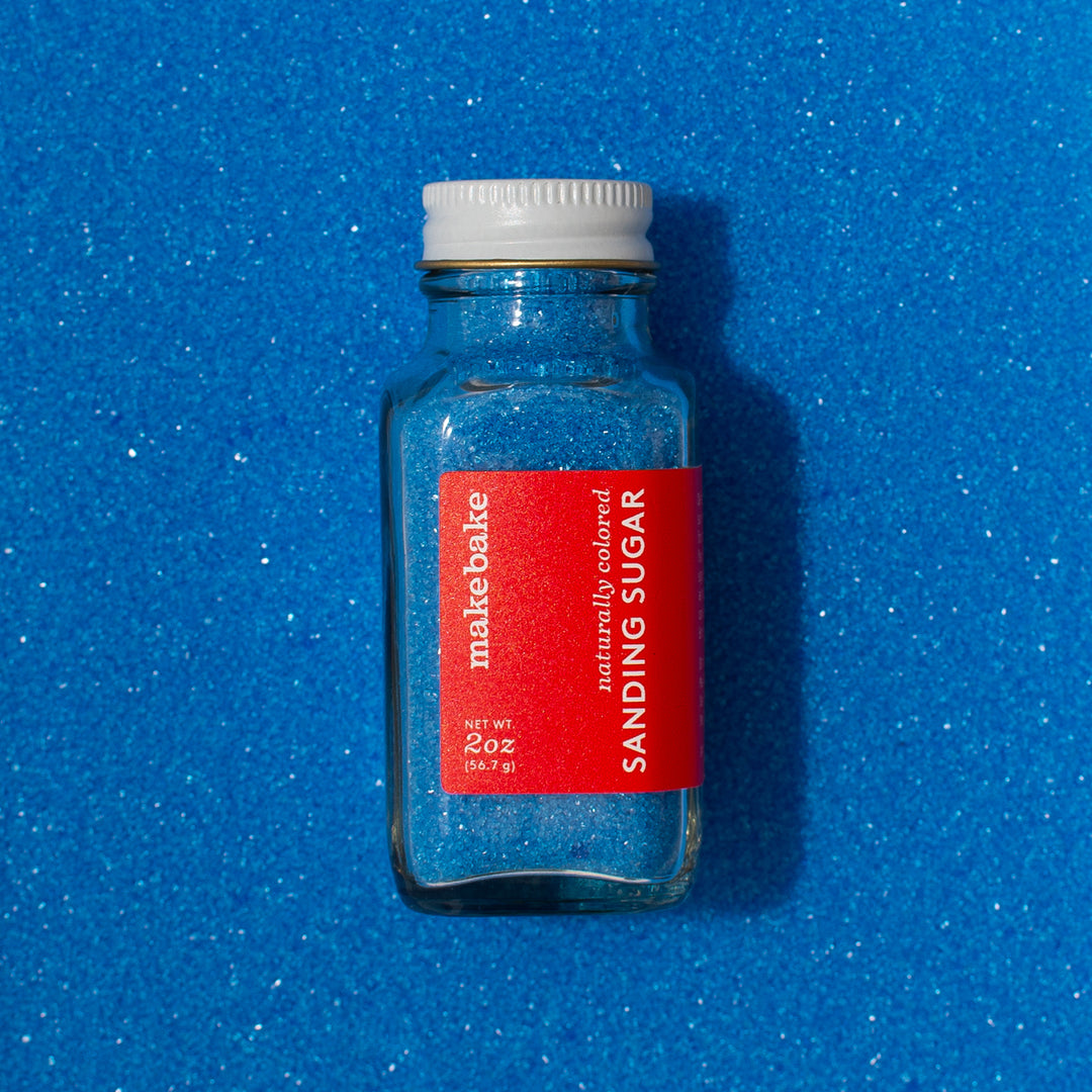 A bottle of Make Bake natural blue sanding sugar with a red label, placed on a dye free blue sanding sugar background.