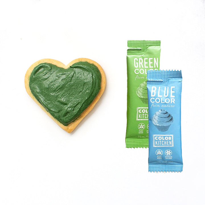 A heart-shaped cookie with forest green frosting next to two packets of food coloring labeled 'GREEN' and 'BLUE' which are combined to make the darker forest colored green food color.