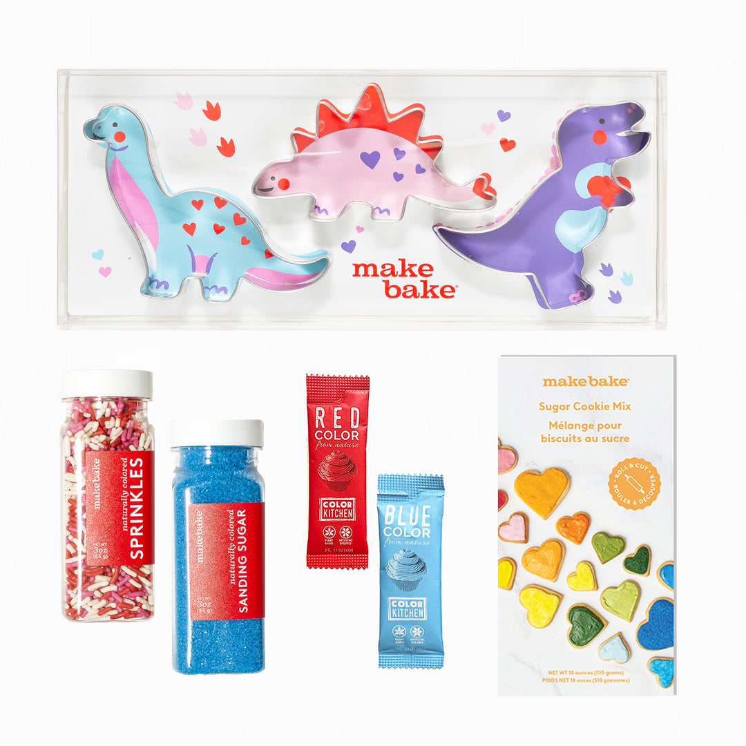 Make Bake valentines dinosaur-themed baking set with colorful dino-shaped cookie cutters, dye free sprinkles and blue sanding sugars and food colors and a box of sugar cookie mix on a white background.