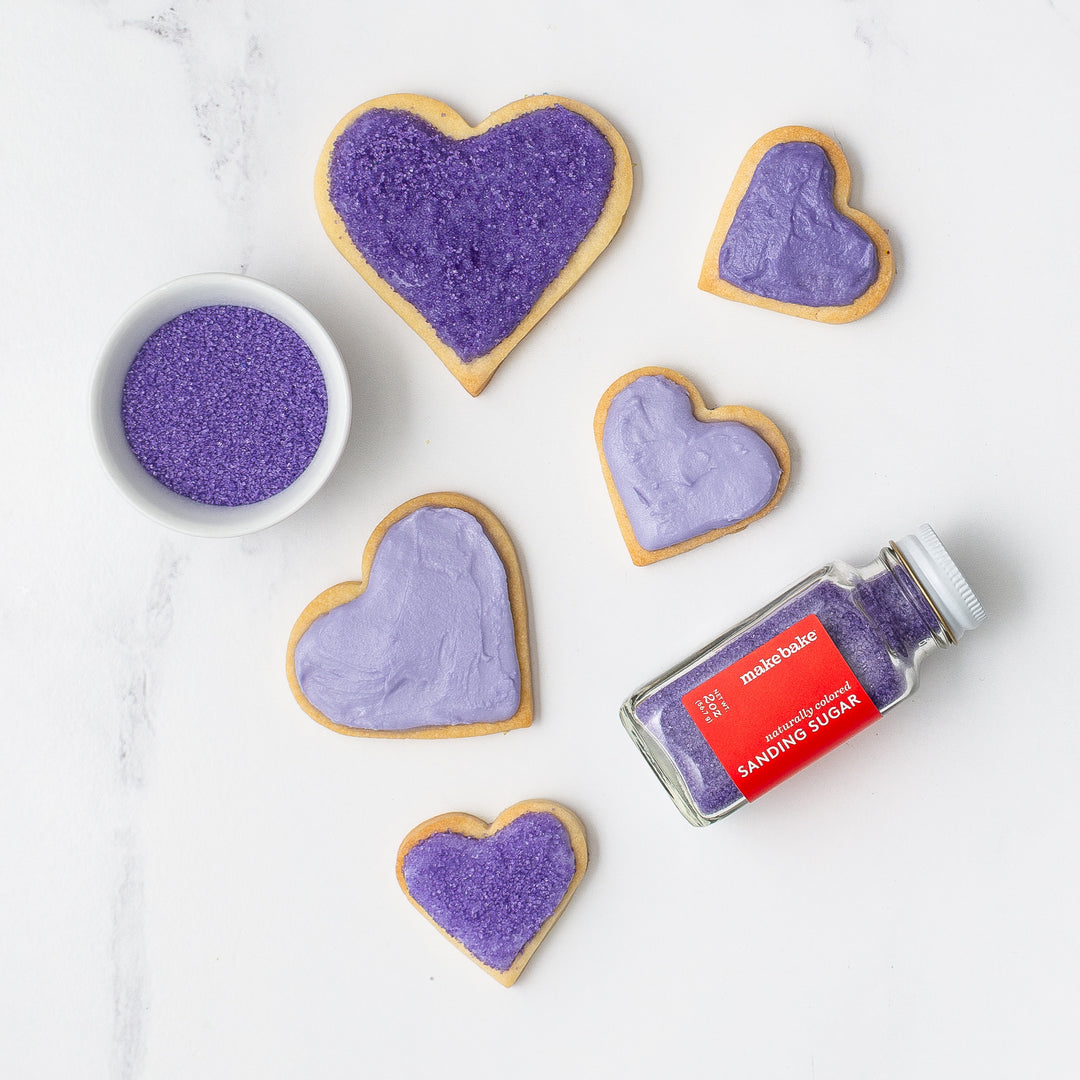 Heart-shaped cookies with purple frosting and a container of purple sanding sugar on a white marble surface.