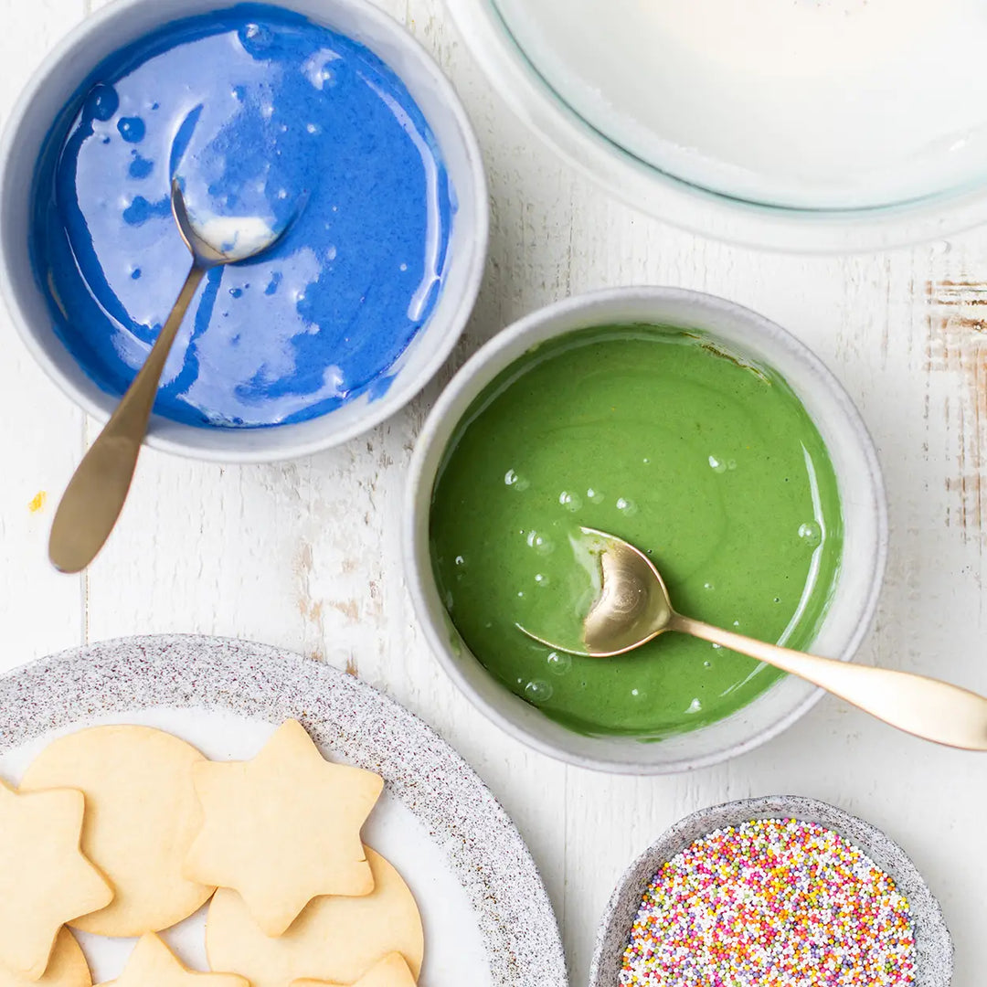 bowls of dye free blue and green icing and undecorated sugar cookies and a bowl of sprinkles on white countertop