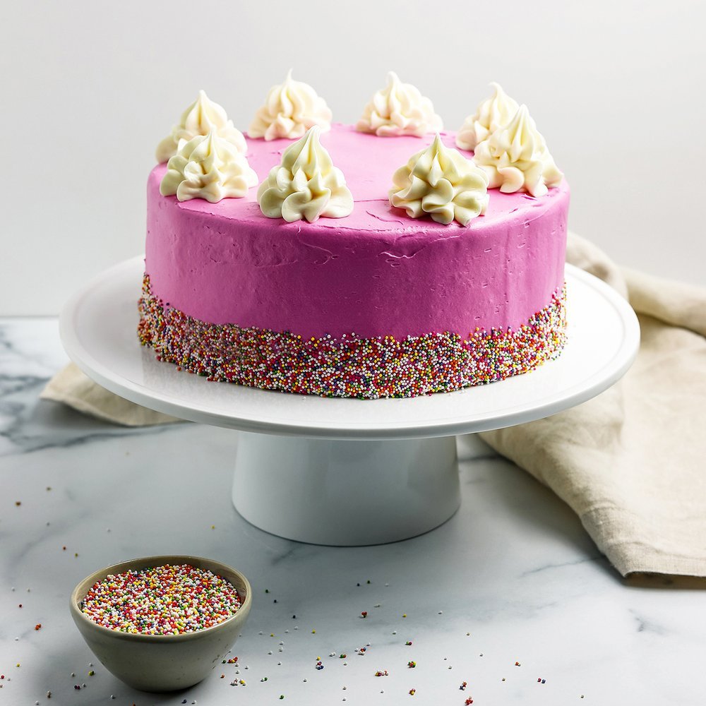 picture of a pink birthday cake on a white cake stand with a bowl of dye free nonpareil sprinkles next to it