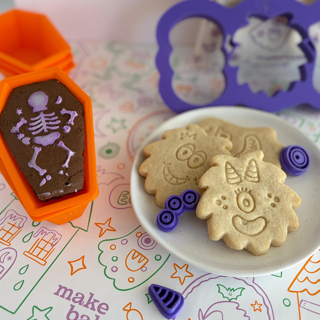 Picture of stamped monster cookies and a coffin shaped chocolate cake with a skeleton stenciled on using frosting in an orange baking cup on a colorful monster printed table runner.