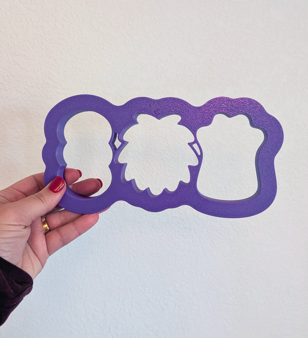 Hand holding a purple cookie cutter that contains three monster shaped heads on a white background.