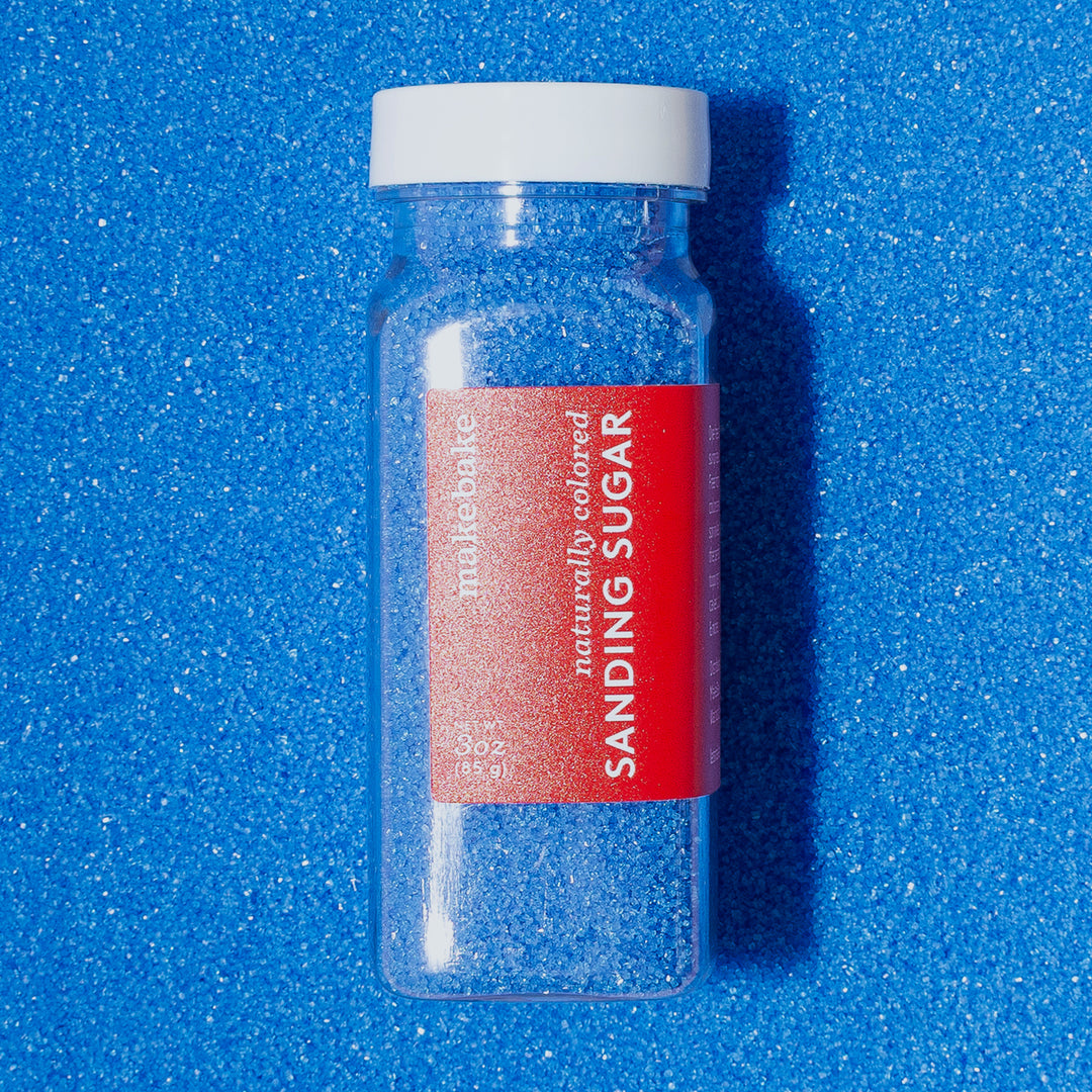 A bottle of Make Bake natural blue sanding sugar with a red label, placed on a dye free blue sanding sugar background.