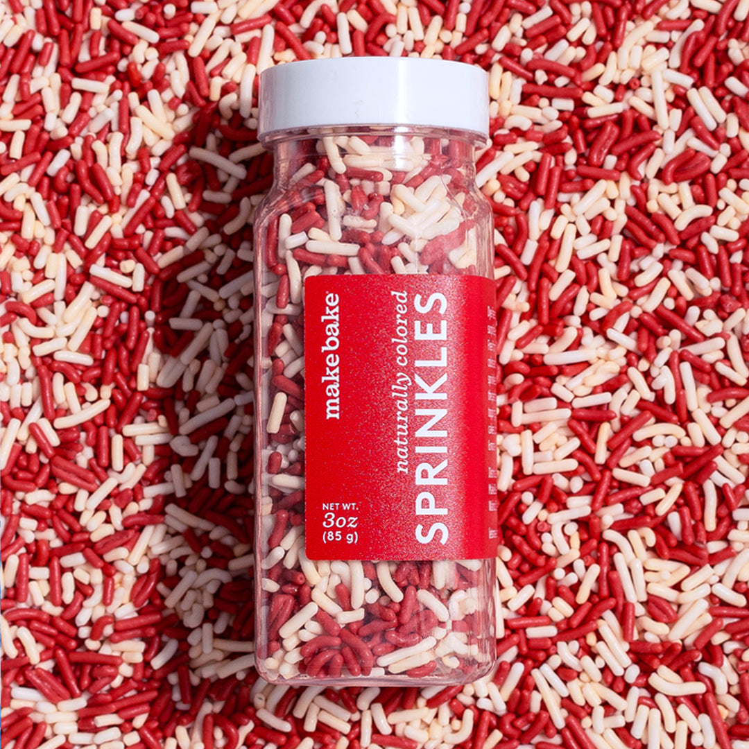 Bottle of dye-free candy cane christmas sprinkles with the red Make Bake label laying on a bed of red and white sprinkles.
