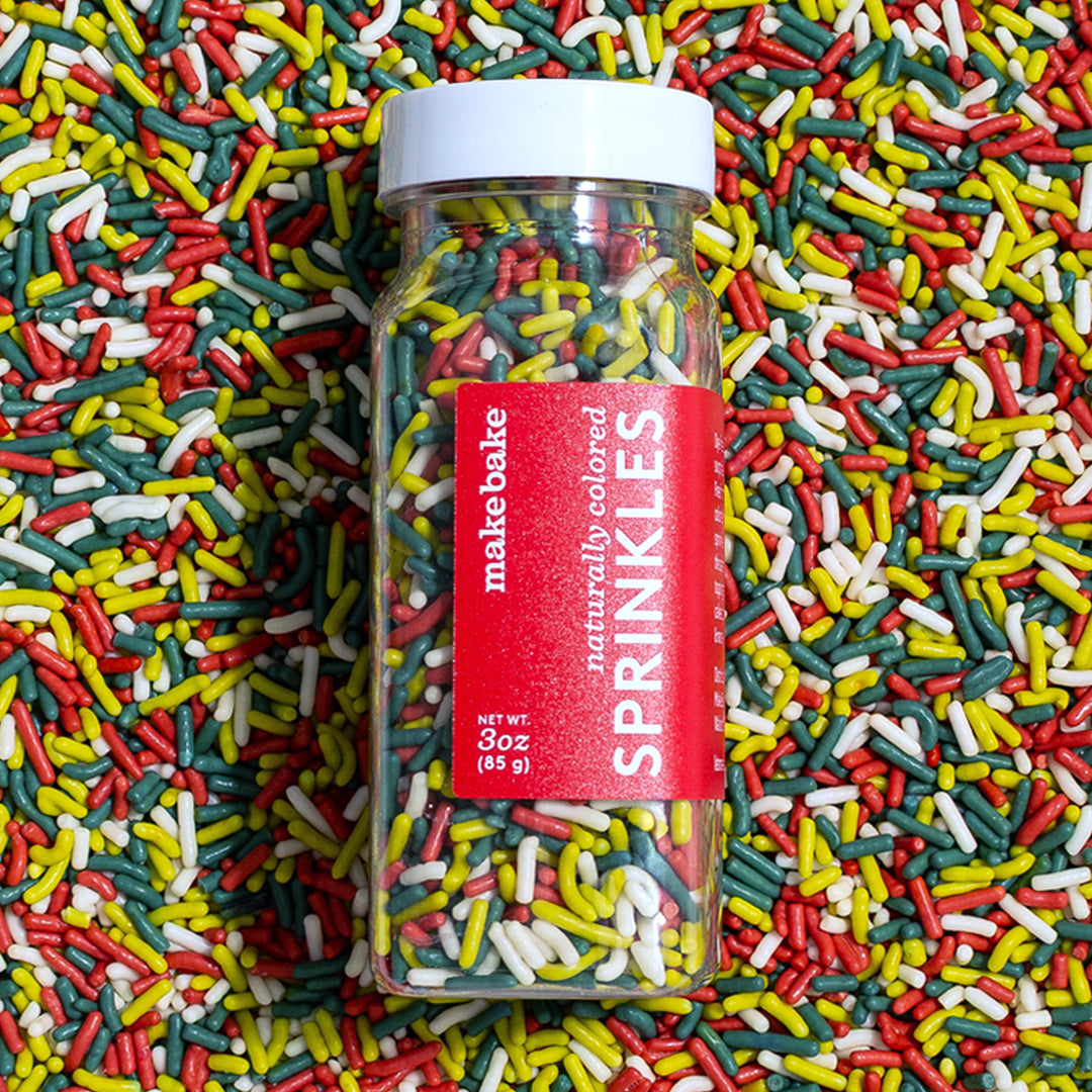 Make Bake brand dye free Christmas sprinkles in red, green and white in the bottle which is laid on top of a bed of the same naturally colored Christmas sprinkles