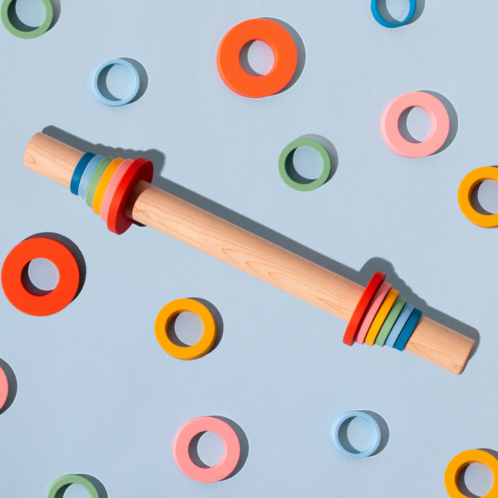 Wooden rolling pin with silicone thickness rings in rainbow colors shown on a blue background.