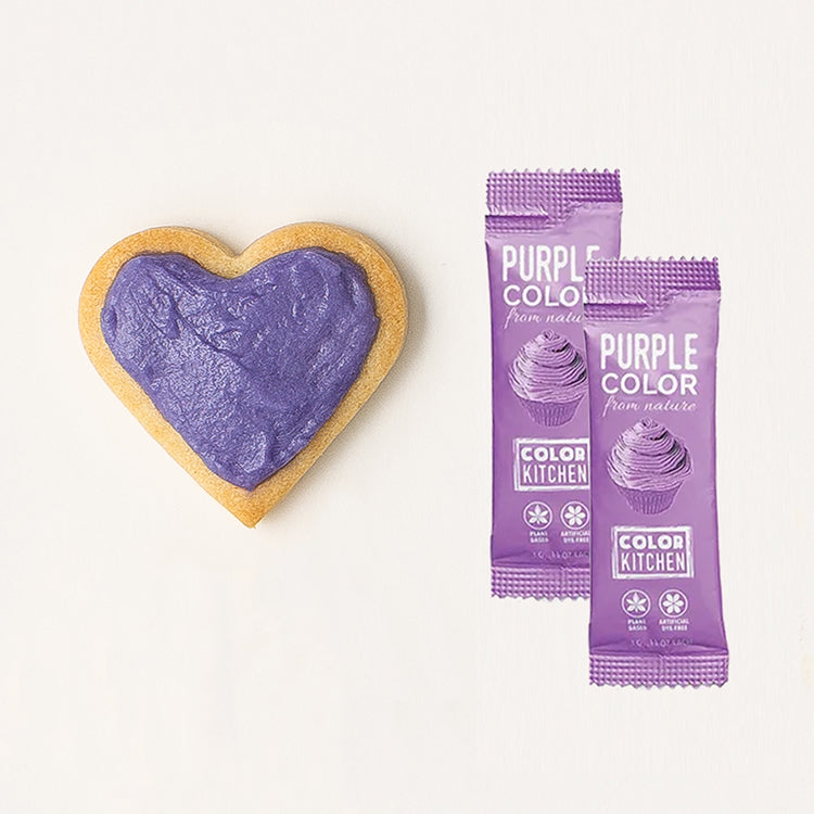 Heart-shaped cookie with purple frosting and two packets of Color Kitchen purple food color on a white background