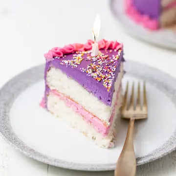 a slice of white birthday cake decorated with dye-free pink and purple frosting, sprinkles and a candle on a white plate