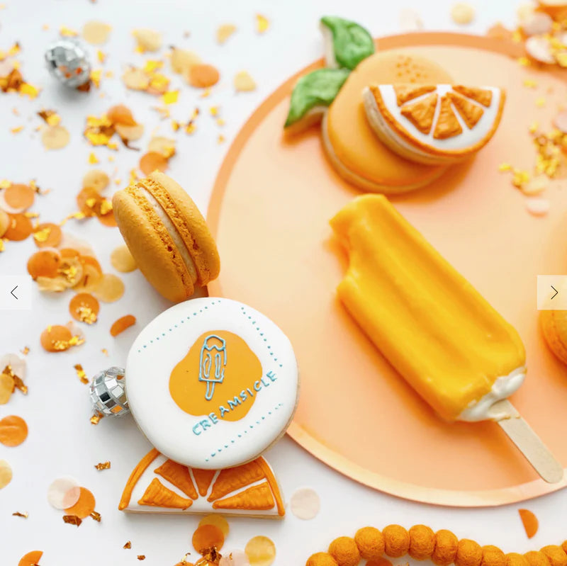 Orange-themed dessert set with cake, macaron, and popsicle and paper orange confetti on a white background.