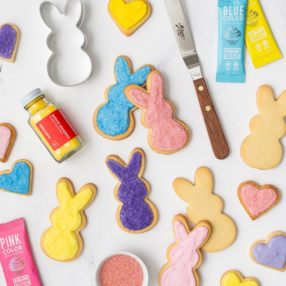 A variety of colorful sugar cookies shaped like bunnies with cookie decorating supplies including a cutter, icing, and sanding sugars visible.