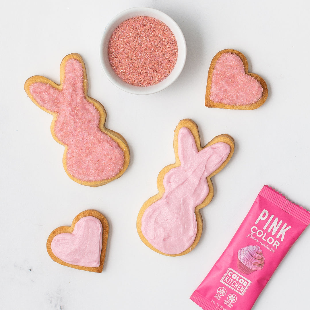 Sugar cookies shaped like bunnies and hearts decorated with dye-free pink sanding sugar and natural pink food colored frosting on a white surface.