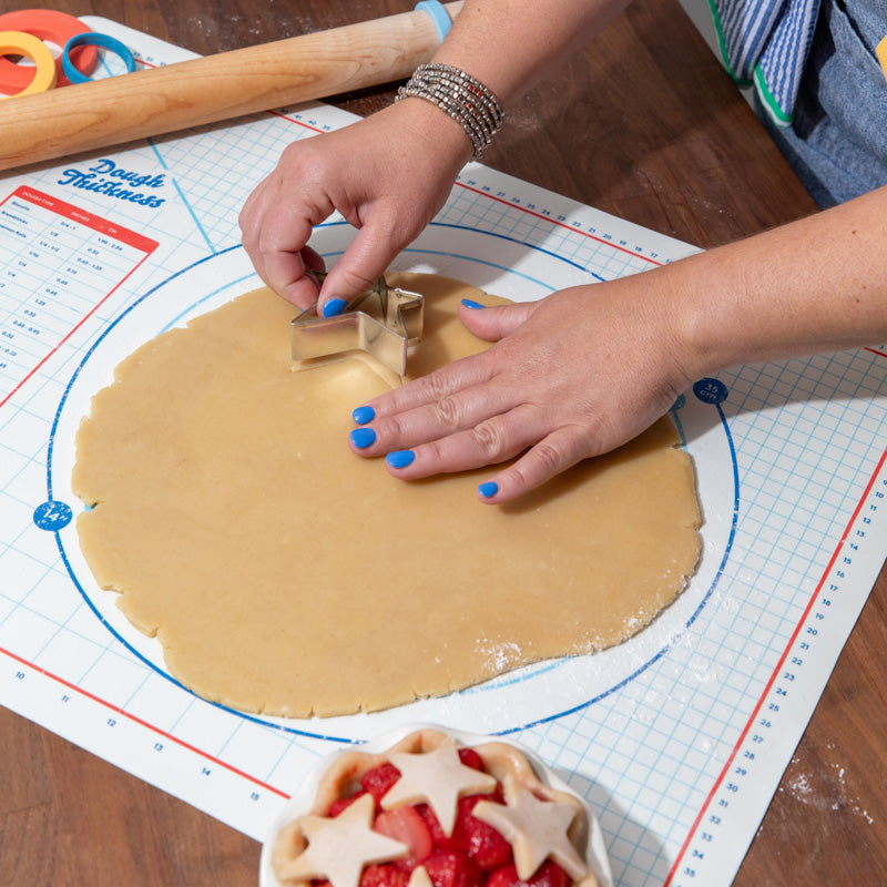 Person cutting out star shaped pie crust from rolled-out dough with a cookie cutter on a Make Bake brand baking mat.