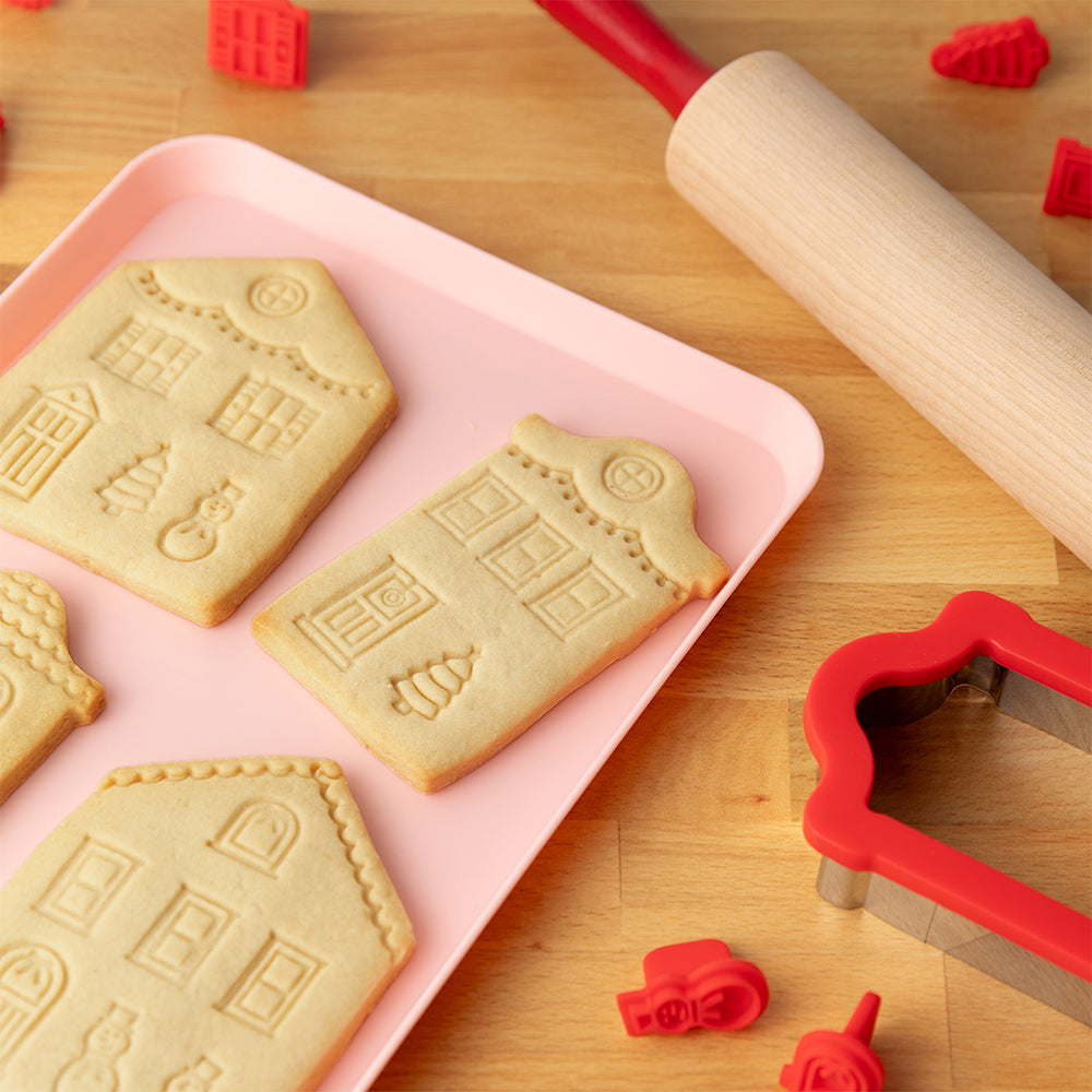 Baked cookies with house shapes on a pink tray, surrounded by cookie cutters and a rolling pin on a wooden surface.
