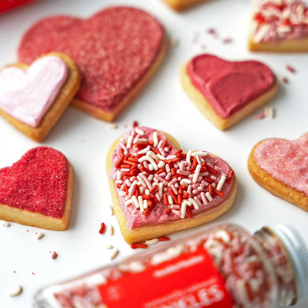 Heart-shaped cookies with red & pink icing and sprinkles on a white surface, with a jar of sprinkles in the foreground.