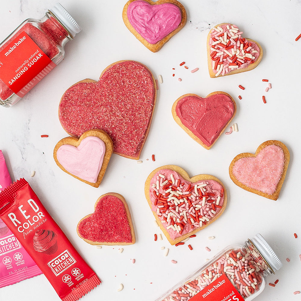 Heart-shaped sugar cookies decorated with pink & red icing, surrounded by dye-free food colors and sprinkles on a white background