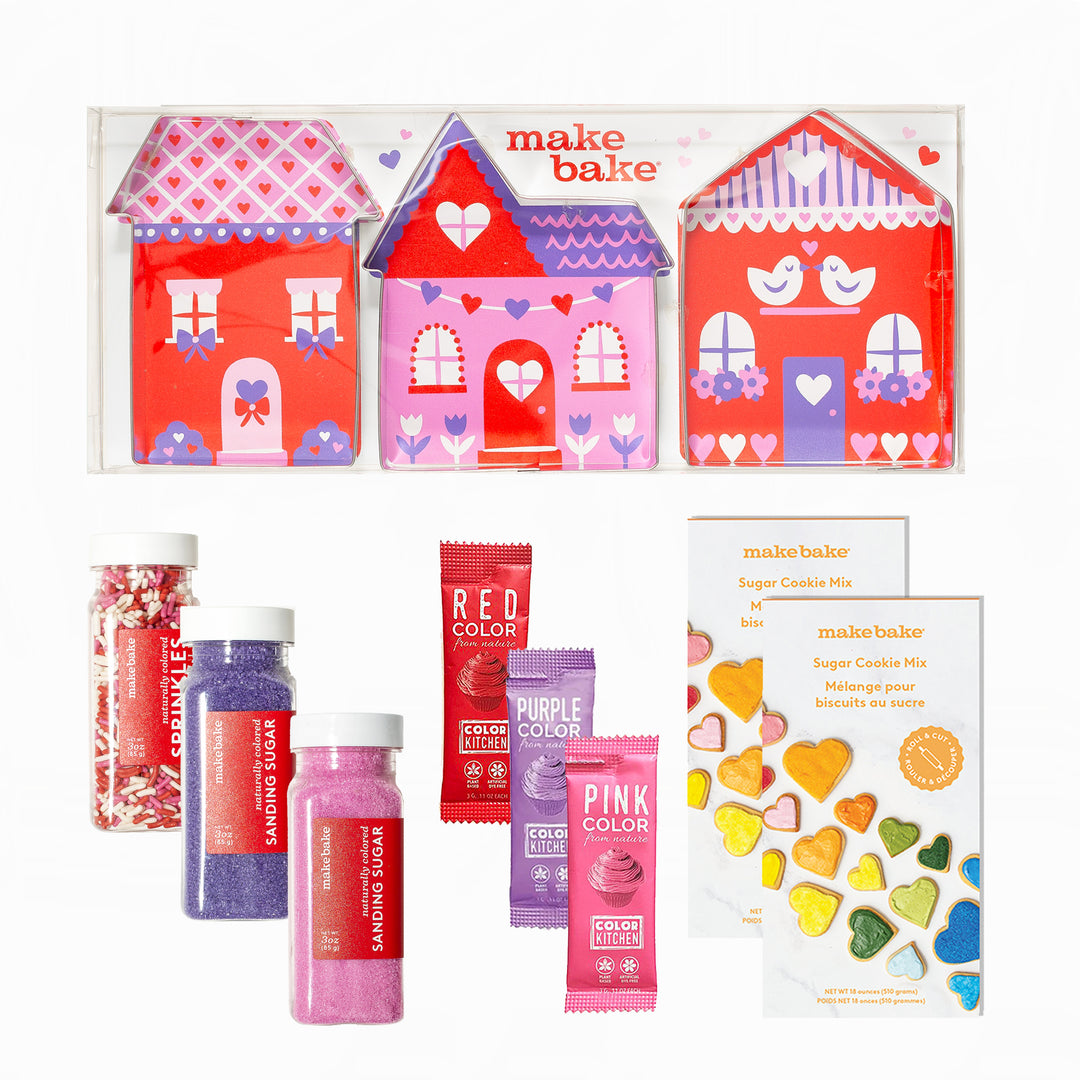 Make Bake cookie making kit with house-shaped cookie cutters and dye free sprinkles and sanding sugars and food colors in red, purple and pink as well as 2 boxes of natural sugar cookie mix on a white background.