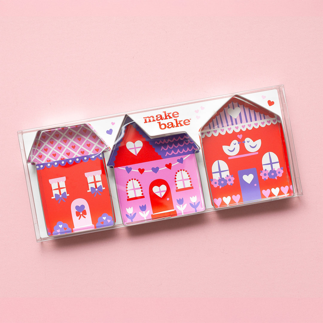 A Make Bake set of three house shaped cookie cutters with illustrated designs in the packaging depicting valentine's themed decorated house in a clear plastic box on a pink background.