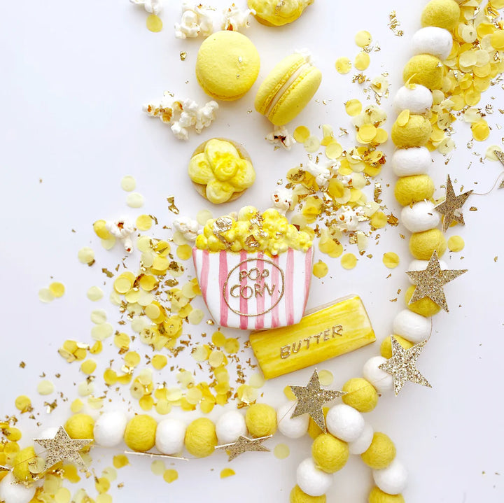 Decorative arrangement with yellow and white macarons, popcorn container, and candy on a white background decorated with yellow confetti.